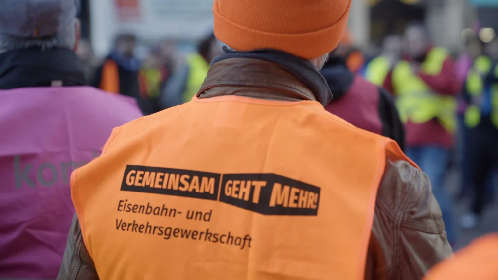 Auf einer Streik-Kundgebung. Ein Mensch trägt eine orangene Verste auf der steht: 