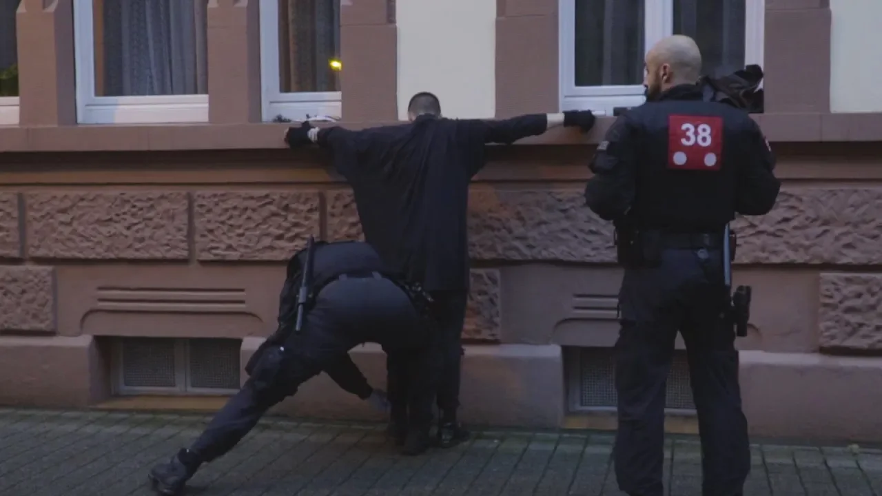 Eine Person wird von zwei Polizisten festgehalten. Sie steht mit ausgestreckten Armen vor einer Hauswand und wird von einem der Polizisten abgetastet.